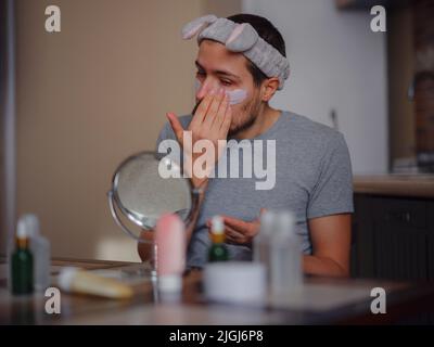 Homme de beauté concept. Caucasien gars vérifiant sa peau. Homme regardant dans le petit miroir tout en appliquant le masque de désintoxication de peau d'argile. Soin de la peau et soin masculin caucasien. Banque D'Images