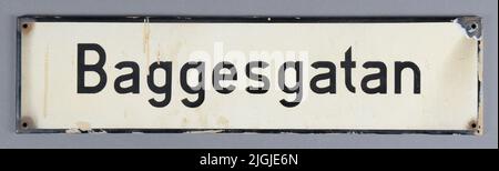 Panneau de Gatoskylt, panneau de rue de tôle. Rectangulaire, émaillé. Blanc avec cadre noir et texte « Baggesgatan ». Quatre trous pour le montage.peinture beige sur les bords de la peinture de façade. L'émail est endommagé dans un coin. Ajouté au 1966-11-18, lors de la démolition de la propriété Varvsgatan 33, kV Drottninghall 11, Karlskrona. Banque D'Images