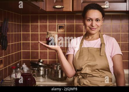 Agréable confiserie charmante femme du Moyen-Orient, femme au foyer en tablier de chef tient un pot de confiture maison sur sa main paume et sourires, regardant Banque D'Images