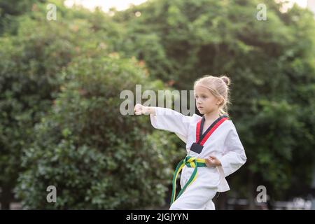 Petite fille caucasienne de sept ans dans un kimono avec ceinture verte jaune faisant de l'exercice de Taekwondo au parc d'été seul pendant le verrouillage du coronavirus covid-19 Banque D'Images