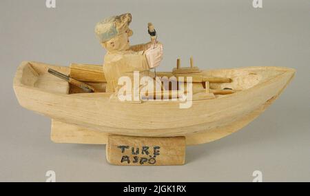 Modèle Modell « Fisherman in boat ». Découpe d'un morceau de bois. Quatre tables, deux planches de siège, deux paires d'années, deux années. Pêcheurs en position assise, coupés en une seule pièce, tenant à Pilkvinda. Sur les récifs est un poisson. Dans le bateau est un vète et un fusil. La casquette du pêcheur, le fusil et le vétérinaire sont peints, le reste non peint. Placez le bateau sous forme de blocs avec une encoche pour le mettre en place. Le texte peint en bleu 'Aspture ö' est alors coupé. Augmenté, 1986-09-30, de ture Petersson, Aspö-Bäck, Drottningskär.Neg n° 86: 148 , 19A Banque D'Images