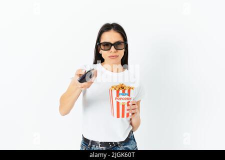 Je ne peux pas choisir quoi regarder, la jeune femme change de chaîne avec la télécommande qui tient le pop-corn et porte 3D lunettes, fond blanc studio Banque D'Images