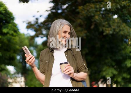 Femme européenne mûre aux cheveux gris heureux de profiter de temps libre après le travail ou de voyager tenant le téléphone tout en prenant un café sur la route à l'aide d'une tasse en papier dans le jardin de la ville ou le parc. Profiter de la vie femme mûre. Banque D'Images