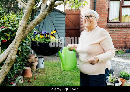 Portrait d'une femme mûre et heureuse, souriante, arrosoir des fleurs avec arrosoir dans le jardin par une journée ensoleillée. Femme âgée à la retraite et jardinage h Banque D'Images