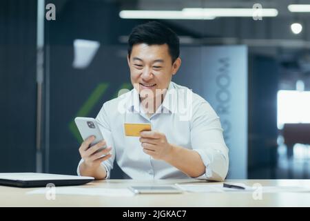 Homme d'affaires asiatique prospère au bureau fait des achats en ligne dans un magasin en ligne, l'homme tient un téléphone et une carte de crédit bancaire dans ses mains Banque D'Images