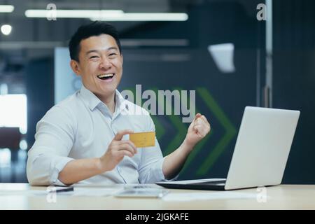 Homme d'affaires asiatique souriant et regardant l'appareil photo, tenant la carte de crédit bancaire pour l'achat dans la boutique en ligne, célébrant la réussite des transactions bancaires, homme au travail Banque D'Images