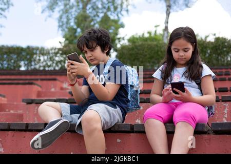 Deux élèves de l'école primaire du caucase utilisant des téléphones mobiles à l'extérieur. Concept de l'utilisation de la technologie à l'âge des enfants. Banque D'Images