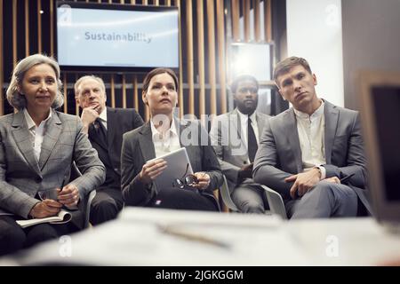 Groupe d'hommes d'affaires multiethniques sérieux en tenue de cérémonie assis dans la salle de conférence et à l'écoute attentive de l'orateur Banque D'Images