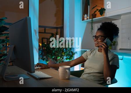 Femme noire positive dans les lunettes parlant sur un smartphone, travaillant sur un pc de bureau tard dans la nuit Banque D'Images