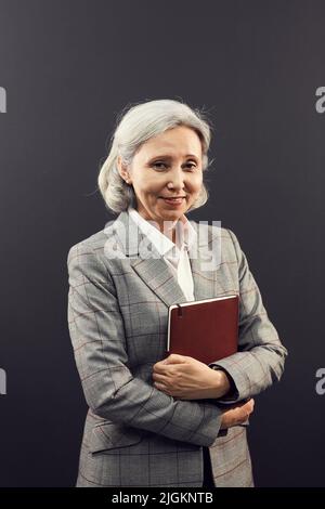 Portrait d'une femme d'affaires asiatique agée et gaie avec des cheveux gris debout sur un arrière-plan isolé et embrassant un organisateur personnel Banque D'Images