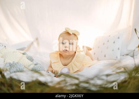 Mignon petite fille en bonne santé dans une robe jaune lying sur l'herbe verte dans le parc sur couverture blanche avec tente, sourire, jouer avec le jouet pour les enfants, développement précoce. Banque D'Images