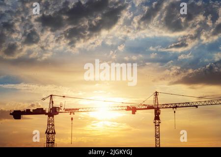 Vue panoramique de deux grues à tour haute silhouette travaillant au début du lever du soleil sur le chantier de construction contre lumière orange vif du soleil ciel nuageux. Ville Banque D'Images