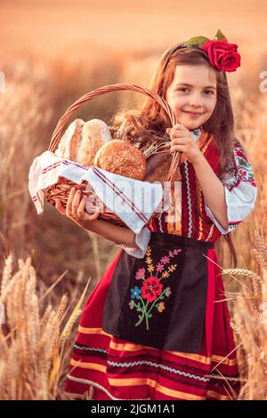 Une femme bulgare ou une jeune fille en robe traditionnelle folklorique tient dans les mains du blé doré et du pain maison fraîchement cuit dans un sac Banque D'Images