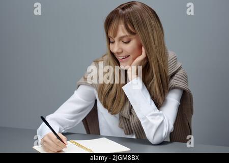 Heureuse jeune blonde étudiante journaliste dame en chaud chandail écriture amour lettre article essai assis sur le lieu de travail dans gris moderne bureau à la maison Banque D'Images