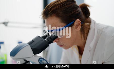 Femme hispanique d'âge moyen portant l'uniforme scientifique au microscope en laboratoire Banque D'Images