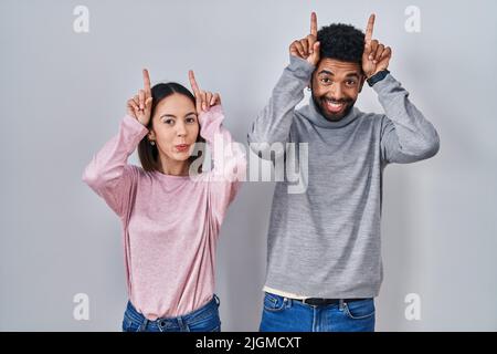 Jeune couple hispanique debout ensemble faisant un geste drôle avec le doigt au-dessus de la tête comme cornes de taureau Banque D'Images