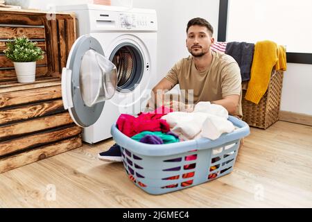 Jeune homme beau mettant le linge sale dans le lave-linge détendu avec une expression sérieuse sur le visage. Simple et naturel regarder l'appareil photo. Banque D'Images