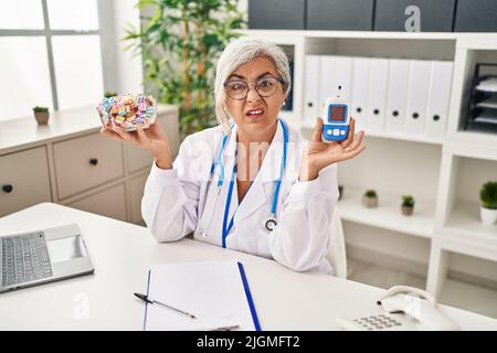 Femme d'âge moyen avec cheveux gris portant l'uniforme de médecin tenant le moniteur de glucose expression sans indice et confuse. Notion de doute. Banque D'Images