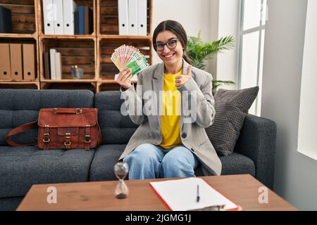 Jeune femme hispanique travaillant au bureau de consultation tenant l'argent souriant heureux et positif, pouce vers le haut faisant l'excellent et signe d'approbation Banque D'Images