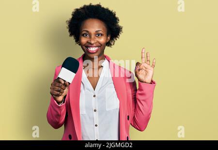 Femme afro-américaine avec cheveux afro tenant le microphone rapporteur faisant signe ok avec les doigts, souriant sympathique gestante excellent symbole Banque D'Images