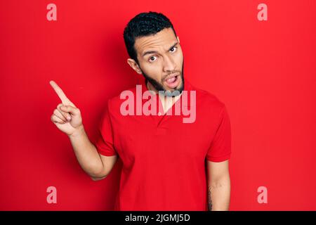 Homme hispanique avec barbe pointant avec les doigts sur le côté dans le visage de choc, regardant sceptique et sarcastique, surpris par la bouche ouverte Banque D'Images