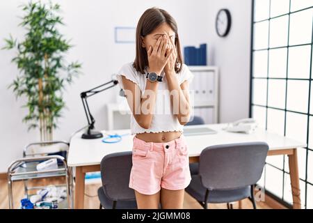 Jeune fille hispanique debout à la clinique pédiatre frottant les yeux pour la fatigue et les maux de tête, l'expression endormi et fatigué. Problème de vision Banque D'Images