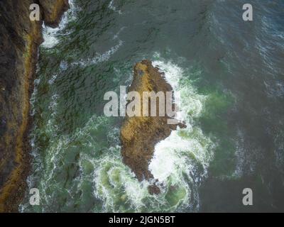 Prise de vue aérienne. Paysage marin. Dans l'eau turquoise claire de l'océan se trouve une grande pierre, un rocher. Des vagues de mousse blanche se brisent sur la côte rocheuse. Banque D'Images