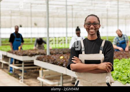 Portrait de femme souriante en serre avec environnement hydroponique avec des travailleurs cultivant des cultures biologiques et des légumes. Agriculteur afro-américain posant heureux devant les rangs avec des semis. Banque D'Images