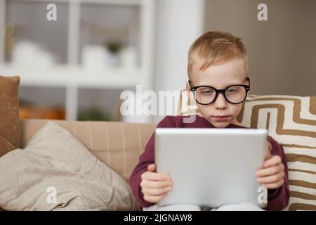 Petit garçon sérieux curieux dans des lunettes assis sur le canapé et en utilisant la tablette numérique tout en lisant le livre en ligne Banque D'Images