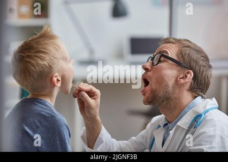 Jeune pédiatre expressif avec le chaume en utilisant le dépresseur de langue et demandant au garçon d'ouvrir la bouche largement pendant les examens médicaux Banque D'Images