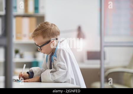 Petit garçon sérieux concentré dans les médecins se vêtir debout à table et de prendre des notes dans le dossier médical Banque D'Images