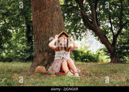 Jolie fille d'école dans un chapeau de paille et la robe est assise sur l'herbe par un arbre et fait une maison d'un livre sur sa tête. Vacances scolaires. Le con Banque D'Images