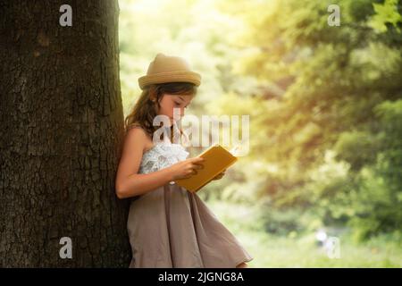 L'été. Une belle fille d'école dans un chapeau de paille et la robe est debout près d'un arbre et de la lecture d'un livre. Vacances scolaires. Le concept de l'extracurrique Banque D'Images