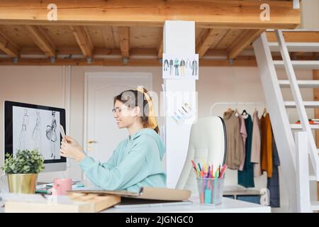 Créateur de vêtements hipster souriant avec queue de cheval foulard, assis au bureau en studio et travaillant sur des croquis de mode Banque D'Images