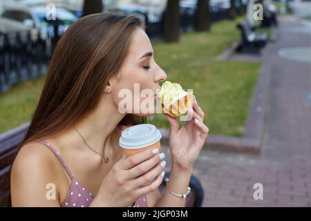 belle fille avec de longs cheveux tient un gâteau dans ses mains et le lèche avec sa langue. séance photo dans la ville. Banque D'Images