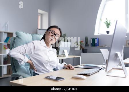 Jeune femme d'affaires asiatique portant des lunettes se sentant épuisée au travail. Tient le dos avec deux mains, se moque, ressent de la douleur et de la fatigue. S'asseoir au bureau à l'ordinateur, a besoin d'une pause. Banque D'Images
