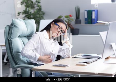 Douleur abdominale sur le lieu de travail. Une jeune belle femme asiatique en lunettes travaille dans le bureau à l'ordinateur, sent la douleur dans son estomac, tient sa main à son estomac, grimace dans la douleur. Banque D'Images