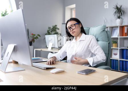 Douleur abdominale sur le lieu de travail. Une jeune belle femme asiatique en lunettes travaille dans le bureau à l'ordinateur, sent la douleur dans son estomac, tient sa main à son estomac, grimace dans la douleur. Banque D'Images