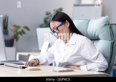 Gros plan. Douleur abdominale sur le lieu de travail.Une jeune femme asiatique en lunettes travaille dans le bureau à l'ordinateur, sent la douleur dans son estomac, tient sa main à son estomac, grimace dans la douleur. Banque D'Images