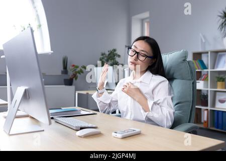 Chaleur sur le lieu de travail. Une jeune femme asiatique assise au bureau, agitant la main, elle fait très chaud, elle se sent mal, elle a besoin d'air frais, faire une pause. Banque D'Images