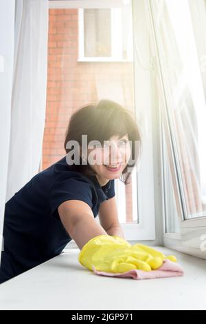 Belle femme avec un sourire balaye la fenêtre de la poussière un jour d'été. Banque D'Images