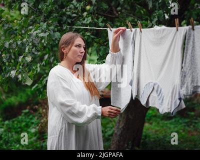 femme caucasienne d'âge moyen en robe blanche faisant des devoirs, pendant des vêtements sur la corde à linge dans la rue dans la cour de la maison de village, concept de jour de blanchisserie Banque D'Images