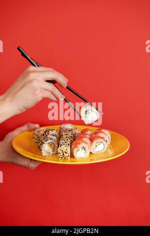 Gros plan une photo de la main femelle tient le rouleau de sushi makizushi servi sur une assiette cuisine japonaise traditionnelle isolée sur fond rouge dans le studio. Copier Banque D'Images