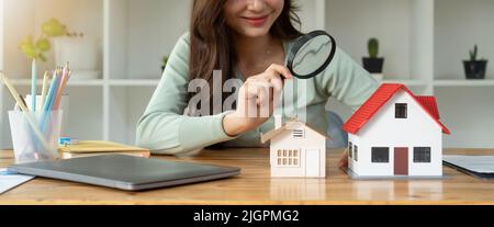 Recherche d'agence immobilière, d'assurance de biens, de prêt hypothécaire ou de maison neuve. Femme avec loupe sur une maison en bois à son bureau Banque D'Images