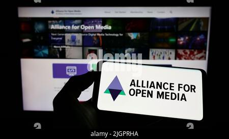 Personne tenant un téléphone portable avec le logo du consortium Alliance for Open Media (AOMedia) à l'écran en face de la page Web d'affaires. Mise au point sur l'affichage du téléphone. Banque D'Images