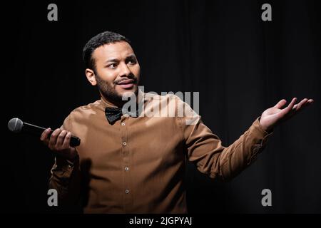 comique indien confus en chemise et noeud papillon tenant le microphone et montrant le geste haussement d'épaules pendant le monologue sur noir Banque D'Images