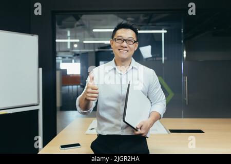 Portrait d'un professeur asiatique réussi, homme dans une salle de classe moderne, regardant l'appareil photo et souriant, portant des lunettes tenant un ordinateur portable, montrant le pouce vers le haut geste affirmatif Banque D'Images