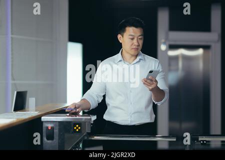 Un homme d'affaires asiatique prospère entre au bureau, utilise la carte-clé pour ouvrir le tourniquet, l'homme regarde l'écran du téléphone lit les nouvelles Banque D'Images
