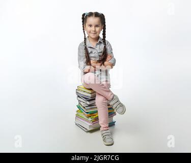 charmante petite fille avec des picots tressés est assise sur une grande pile de livres. Banque D'Images