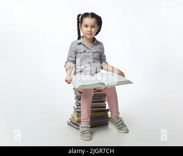 charmante petite fille avec des picots tressés est assise sur une grande pile de livres. Banque D'Images